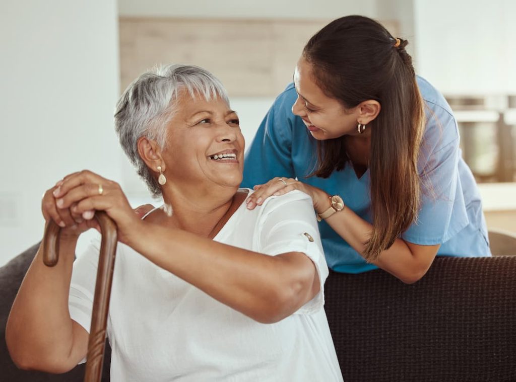 Happy, relax and senior woman with caregiver smile while sitting on a living room sofa
